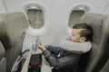 A boy is sitting in a plane near the porthole With travel pillow ,playing in a gadget and waiting for take-off.