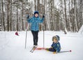 A boy with skis on sits in the snow next to his smiling mother in the winter forest