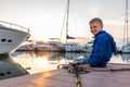 A boy with a smile in a blue jacket sits on a pier with yachts during sunset
