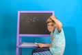 The boy solves the math equation 2 + 2 written on the blackboard. The child scratches his head because he doesn`t know the answer