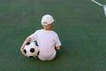 A boy in sports clothes sitting on a green lawn on a football field with a soccer ball back, back view, sports section, training