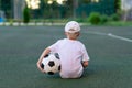 A boy in sports clothes sitting on a green lawn on a football field with a soccer ball back, back view, sports section, training