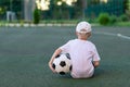 A boy in sports clothes sitting on a green lawn on a football field with a soccer ball back, back view, sports section, training