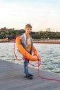 A boy is standing on a pier near the sea and wearing an orange lifebuoy