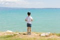 Boy stands on coast of lake, sunny daytime. Healthy leisure. Summer. Calmness. Nature. Panoramic view. Blue clear water