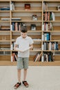 A boy stands in the library and reads a book while standing. Preparing for homework. The boy loves to read. Free space at the Royalty Free Stock Photo