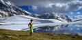 A boy stands next to the mountain lake in front of snow mountains in the sunny and cloudy day