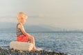 Boy in striped t-shirt sitting on the beach with a stick. Sea, mountains and city with skyscrapers in the distance against the bac Royalty Free Stock Photo