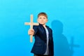 A boy in a suit holds a Catholic cross in front of him against a blue background