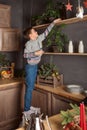 A boy in a sweater and jeans is standing on a stool and trying to reach the upper kitchen shelves in the evening