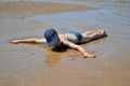 A boy in swimming trunks and a cap lies face down on a sandy beach after a low tide, with his arms spread wide.