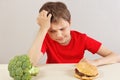 Boy at the table chooses between hamburger and fresh broccoli on white background