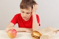 Boy at the table chooses between hamburger and fruit on white background