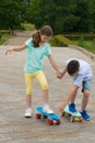 The boy teaches the girl how to ride on a board with wheels in a park on the road