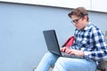 Boy teenager schoolboy or student is sitting on the stairs, working in the computer, wearing glasses, in a shirt, smiling, red b Royalty Free Stock Photo