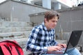 Boy teenager schoolboy or student is sitting on the stairs, working in the computer, wearing glasses, in a shirt, smiling, red b Royalty Free Stock Photo