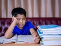 A boy is tired of reading a book on a wooden table. with a pile of books beside The background is a red sofa and cream curtains Royalty Free Stock Photo