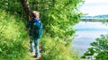 A boy with a tourist backpack walks on a hiking route with trekking poles around a picturesque mountain forest lake. Orienteering