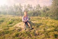 A boy traveler with a backpack and trekking poles is resting on a stone in the fog