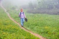 A boy traveler with hiking poles walks along the path on the green slope in heavy fog