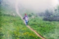 A boy traveler with hiking poles walks along the path on the green slope in heavy fog