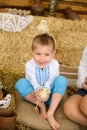 boy in Ukrainian embroidered shirt sits on pile of hay, plays with chickens, one of them is on his head
