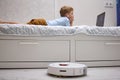 Boy uses a laptop on the bed while the robot vacuum cleaner does the cleaning Royalty Free Stock Photo