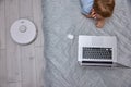 Boy uses a laptop on the bed while the robot vacuum cleaner does the cleaning Royalty Free Stock Photo