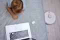 Boy uses a laptop on the bed while the robot vacuum cleaner does the cleaning Royalty Free Stock Photo