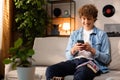 A boy uses a smartphone while teaching English at home. A young man wearing a denim shirt sits on a couch in the living