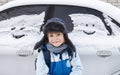 boy in a warm fur hat stands next to a car covered with snow and human faces painted on the windows