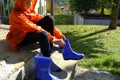 Boy wearing orange raincoat putting on blue rubby boots sitting on stairs in backyard. Clothing for rainy weather