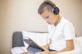 A boy in white T-shirt on the bed at home playing with pc tablet or reading online and listening to music with headphones Royalty Free Stock Photo