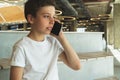 Boy in white t-shirt is sitting indoors and talking on his mobile phone. A teenager uses a cell phone, calling, phoning Royalty Free Stock Photo