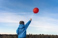 A boy in a winter blue jacket and hat stands with a red balloon on a stick