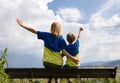 boy and woman in blue and yellow t-shirts sit with their backs against sky raising hands joyfully Royalty Free Stock Photo