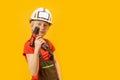 Boy 9-10 years old imitates builder, isolation on yellow background. Child wears helmet and working overalls holds drill in his