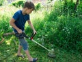 A boy 11 years old mows the grass with an electric scythe on the lawn in the yard of a house on a sunny summer day.