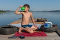 A boy of 11 years old slake his thirst with water from a mug, sitting on a wooden pier, against the backdrop of the lake on a