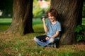 The boy of 8-9 years sits, leaning against tree and holds the tablet on a lap.