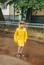 A boy in a yellow raincoat walks outside in the rain. A child jumps on the narrows. jump through muddy puddles.