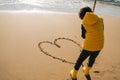 Boy in yellow rubber boots drawing heart shape on sand at the beach. School kid touching water at autumn winter sea Royalty Free Stock Photo