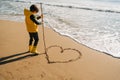 Boy in yellow rubber boots drawing heart shape on sand at the beach. School kid touching water at autumn winter sea Royalty Free Stock Photo