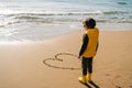 Boy in yellow rubber boots drawing heart shape on sand at the beach. School kid touching water at autumn winter sea Royalty Free Stock Photo