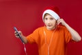 A boy in a yellow T-shirt and a red Santa hat holds a phone in his hands and listens to music through headphones Royalty Free Stock Photo