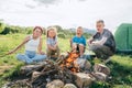 Boys and girls kids cheerfully laughing and roasting marshmallows and sausages on sticks over campfire flame near the green tent. Royalty Free Stock Photo