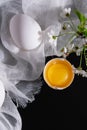 A branch of blooming cherry and a whole and broken egg on white gauze and a black background. Easter