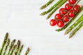 A branch of fresh cherry tomatoes and some asparagus stems in two opposite corners of the off-white rustic table.