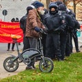 Older man with white beard and blue knitted cap pushes a tiny electric folding bike in front of a group of police officers in