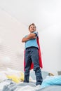 Brave proud little boy in a superhero costume stands on the bed, imagining flying